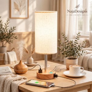 May include: A cylindrical table lamp with a beige fabric shade and a light wood base featuring a silver metal stem. The base includes two USB ports and a touch-sensitive power button. A smartphone is connected to the lamp via a black charging cable.