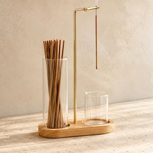 Peut inclure: Un ensemble porte-encens composé d'une base en bois clair avec deux cylindres en verre. Un cylindre haut contient un faisceau de bâtonnets d'encens bruns, tandis qu'un bras en métal doré suspend un bâtonnet au-dessus d'un cylindre plus court.