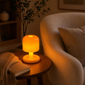 May include: A portable mushroom-shaped table lamp with a smooth, translucent orange body that emits a warm, glowing light. The lamp features a rounded dome shade, a cylindrical stem, and a circular base, creating a soft, ambient illumination.