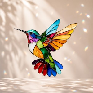 Puede incluir: Un atrapasoles de colibrí hecho de vidrieras con un mosaico vibrante de colores que incluye azul, verde, amarillo, naranja, rojo, morado y blanco. El ave se muestra en vuelo con las alas extendidas y un pico largo, fino y negro.