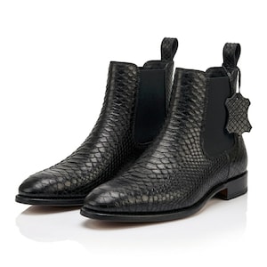 Op de afbeelding: Een paar zwarte Chelsea boots met een textuur van slangenleer, elastische zijpanelen, een treklipje bij de hiel en een klein bijpassend leren label aan de zijkant. De laarzen hebben een puntige neus en een ontwerp met een lage, gestapelde hak.