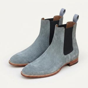 Op de afbeelding: Een paar lichtblauwe suède Chelsea boots met zwarte elastische zijpanelen, een ronde neus, een lage houten hak en een lichtblauw treklipje aan de achterkant van elke enkel. De laarzen hebben een gladde textuur en een natuurlijke bruine leren zoolrand.