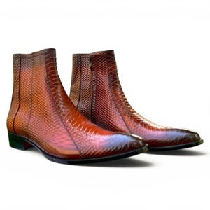 May include: A pair of ankle boots featuring a textured, snakeskin-patterned leather finish in a gradient of deep brown and reddish-orange tones. The boots have a pointed toe, a side zip closure, and a low, stacked heel with a dark sole.
