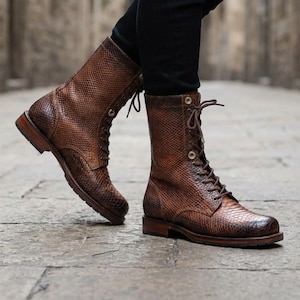 Könnte beinhalten: Ein Paar wadenhohe Schnürstiefel mit strukturiertem braunem Schlangenledermuster. Die Stiefel haben eine abgerundete Zehenpartie, einen Absatz in Holzschichtoptik und dunkelbraune Schnürsenkel, die durch Metallösen bis zur Wadenmitte geführt werden.