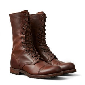 Könnte beinhalten: Ein Paar wadenhohe Schnürstiefel aus sattem braunem Leder. Die Stiefel verfügen über eine abgerundete Zehenpartie mit dekorativen Brogue-Details, einen Schichtabsatz und passende braune Schnürsenkel, die durch Metallösen am Schaft verlaufen.