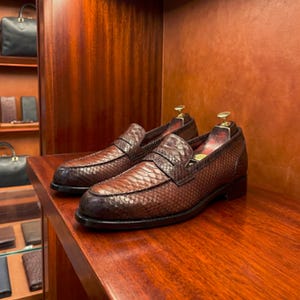 Peut inclure: Une paire de mocassins en peau de serpent marron avec un motif à écailles texturé, un bout arrondi et un petit talon noir. Chaque chaussure contient un embauchoir en métal doré à l'intérieur, posée sur une étagère en bois poli.