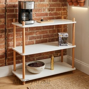 May include: A three-tier shelving unit with white rectangular shelves and light wood cylindrical supports. The top shelf holds a silver and black coffee maker and a small wooden cutting board. The middle shelf holds a boxed figurine. The bottom shelf holds a ceramic bowl filled with coffee beans and a tall brass-colored manual coffee grinder.