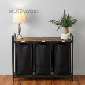 May include: A laundry sorter featuring a black metal frame with a dark wood-toned slatted top surface. It includes three removable black fabric bags with integrated handles, designed for organization. The text MLKByAfzal! is visible.