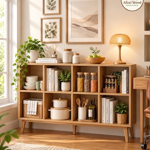 May include: A light wood eight-cube storage shelf with tapered legs, filled with books, white ceramic kitchenware, glass jars of grains, a woven basket, potted plants, and wooden utensils. The logo Afzal Wood Home & Decor is in the corner.