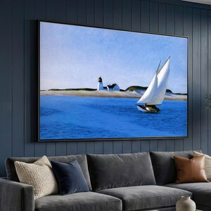 Op de afbeelding: Een rechthoekige ingelijste kunstdruk met een schilderij van een witte zeilboot op felblauw water. Op de achtergrond staan een witte vuurtoren en een klein gebouw op een zandduin onder een heldere, lichtblauwe lucht. Het kunstwerk hangt.