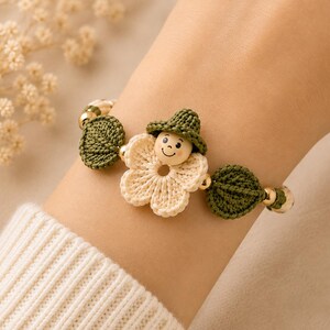 May include: A bracelet featuring a central cream-colored crocheted flower, a small wooden bead with a painted face, and a tiny green crocheted hat. The design includes dark green crocheted leaf accents and small gold-toned metallic beads.