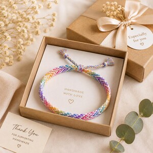 May include: A braided friendship bracelet featuring a multi-colored chevron pattern in pastel shades of pink, blue, yellow, and purple. The bracelet is presented inside a square brown cardboard gift box with a card that reads HANDMADE WITH LOVE.