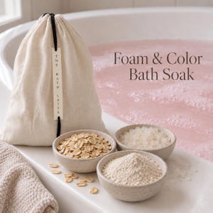 May include: A beige drawstring fabric bag labeled The Bath Latte, accompanied by three small bowls containing rolled oats, white bath salts, and a fine powder. The text Foam & Color Bath Soak is visible above a bathtub filled with pink foamy water.