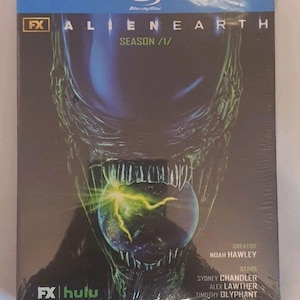 Puede incluir: Una caja de Blu-ray para la temporada 1 de Alien Earth con una criatura alienígena oscura de tonos verdes que sostiene un planeta brillante en su boca. La portada muestra el texto FX, Alien Earth, Season 1, el creador Noah Hawley y las estrellas Sydney Chandler, Alex Lawther y Timothy Olyphant, con los logotipos de FX y Hulu en la parte inferior.