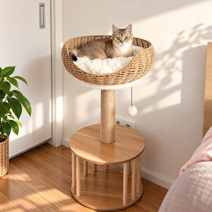 Puede incluir: Un árbol para gatos con una base redonda de madera clara con soportes verticales, un poste central para rascar envuelto en sisal y una cesta superior de ratán trenzado forrada con un cojín blanco afelpado. Un pompón blanco cuelga de la cesta.