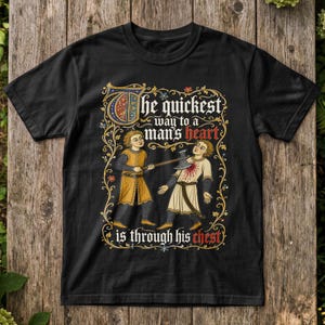 Op de afbeelding: Een zwart t-shirt met een middeleeuwse illustratie van een persoon die een ander met een bijl slaat. De tekst luidt: The quickest way to a man's heart is through his chest. Het ontwerp is omlijst door een gedetailleerde, kleurrijke bloemenrand.