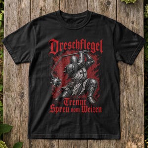 Könnte beinhalten: Ein schwarzes T-Shirt mit einer Grafik eines mittelalterlichen Ritters in Rüstung, der einen Dreschflegel schwingt. Der Text "Dreschflegel" steht über dem Ritter und der Text "Trennt Spreu vom Weizen" ist darunter in einer roten Schrift gedruckt.