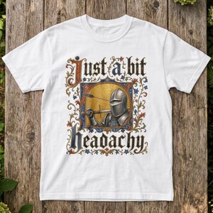 May include: A white short-sleeved t-shirt featuring a graphic of a medieval knight in armour with an arrow through the helmet. The design includes ornate floral borders and the text "Just a bit headachy" in a gothic-style black font.