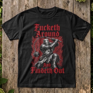Könnte beinhalten: Ein schwarzes Kurzarm-T-Shirt mit einer Grafik eines knienden mittelalterlichen Ritters in Rüstung, der einen Stachelflegel hält. Der Text "Fucketh Around And Findeth Out" ist in roter gotischer Schrift über und unter dem Ritter gedruckt.
