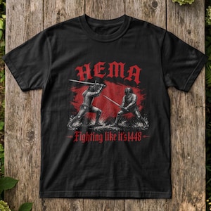 Puede incluir: Una camiseta negra de manga corta con un gráfico rojo de dos figuras con armadura histórica batiéndose en duelo con espadas. El texto HEMA está impreso en rojo sobre las figuras y el texto Fighting like it's 1448 está impreso en rojo debajo.