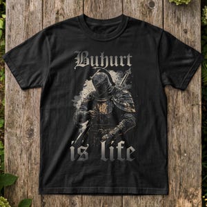 May include: A black short-sleeved t-shirt featuring a graphic print of a medieval knight in full armour holding a sword. The text Buhurt is printed in a gothic font above the knight, and the words is life are printed below in the same font.