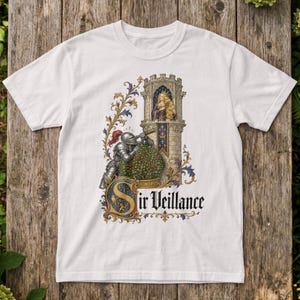 Op de afbeelding: Een wit t-shirt met korte mouwen en een grafische illustratie van een ridder in harnas die door een telescoop naar een persoon in een stenen toren kijkt. Het ontwerp bevat florale versieringen en de tekst Sir Veillance in zwarte letters.