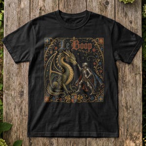May include: A black t-shirt featuring a graphic print of a knight in silver armour touching the nose of a large dragon. The design includes ornate gold and blue floral borders, a small shield emblem at the bottom, and the text Le Boop in a gothic font.