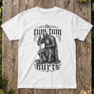 Może przedstawiać: Biały t-shirt z krótkim rękawem z czarną grafiką przedstawiającą rycerza w pełnej zbroi, klęczącego i trzymającego miecz. Napis "the tum tum hurts" jest wydrukowany gotycką czcionką nad i pod rycerzem, w otoczeniu ozdobnych zawijasów.