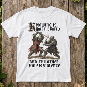 Puede incluir: Una camiseta blanca de manga corta con un gráfico de dos caballeros con armadura metálica luchando con espadas. El texto superior dice "KNOWING IS HALF THE BATTLE" y el texto inferior "AND THE OTHER HALF IS VIOLENCE" en una fuente gótica negra.
