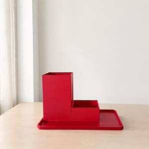 Pode incluir: Um conjunto organizador de secretária vermelho brilhante composto por um porta-canetas retangular alto, uma caixa de armazenamento quadrada mais curta e uma base de tabuleiro retangular plana. As peças formam uma unidade compacta.