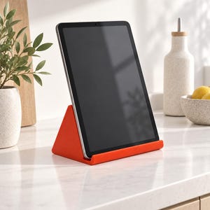 Könnte beinhalten: Ein leuchtend orangefarbener, dreieckiger Tablet-Ständer, der ein dunkelgraues Tablet im Hochformat hält. Der Ständer verfügt über eine breite Basis und eine vertiefte Rille, um das Gerät sicher zu stützen und einen stabilen Blickwinkel zu bieten.