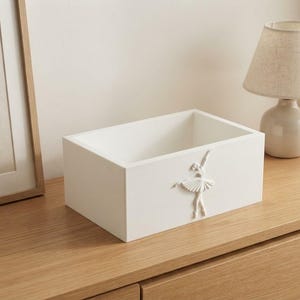 Peut inclure: Une boîte de rangement rectangulaire blanche ornée d'un relief en trois dimensions représentant une ballerine en tutu sur la face avant. La boîte présente une finition lisse et mate ainsi qu'une ouverture sur le dessus.