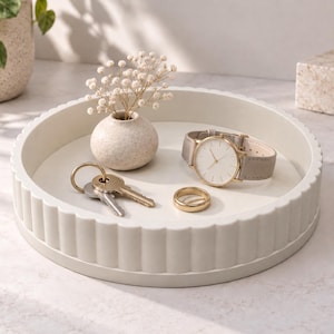 Könnte beinhalten: Ein rundes, cremeweißes Dekotablett mit geriffeltem, gewelltem Rand. Im Tablett befinden sich eine kleine, gesprenkelte beige Vase mit getrockneten weißen Blumen, eine goldfarbene Armbanduhr mit grauem Armband, ein goldener Ring und ein Schlüsselbund.