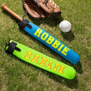Puede incluir: Un bate de béisbol de madera dentro de una funda de neopreno azul con el nombre ROBBIE en letras amarillas, una funda de neopreno verde lima con el nombre CARLIE en letras amarillas con costuras, un guante de cuero y una pelota de béisbol.