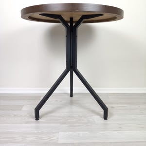 May include: A round table with a dark brown wooden top and a black metal base. The base features three angled legs that meet at a central vertical support column, which connects to the underside of the tabletop with three metal brackets.