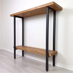 Puede incluir: Una mesa consola con dos estantes horizontales de madera con acabado de veta natural, sostenida por un marco de metal negro que consta de cuatro patas rectangulares verticales conectadas a los estantes en las esquinas con herrajes visibles.