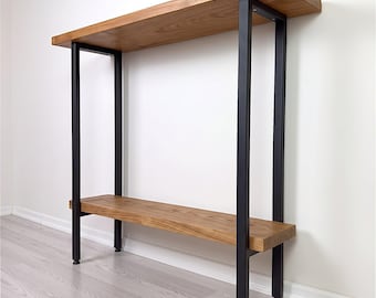 Pode incluir: Uma mesa consola com duas prateleiras horizontais de madeira com acabamento de veio natural, suportada por uma estrutura de metal preto composta por quatro pernas verticais retangulares ligadas às prateleiras nos cantos com ferragens visíveis.
