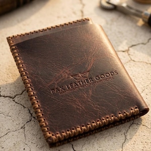 Peut inclure: Un portefeuille en cuir marron foncé avec une surface texturée et des coutures visibles sur les bords. Le devant est gaufré avec le texte PAX LEATHER GOODS HANDMADE IN CONSTANTINOPLE et un petit graphique de bateau en papier au-dessus.