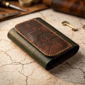 Peut inclure: Un portefeuille en cuir vert foncé doté d'un rabat rectangulaire en cuir marron avec des coutures apparentes. La partie en cuir marron est gravée des noms NIDHAL, MARWA et OUSSAYD disposés verticalement. Le portefeuille a une finition lisse.