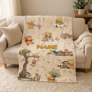 Könnte beinhalten: Eine beige Fleecedecke mit verschiedenen Illustrationen von Winnie Puuh und seinen Freunden, darunter Tigger, I-Aah und Ferkel. Das Design zeigt verstreute Blätter und das Wort NAME in großen orangefarbenen Buchstaben in der Mitte.