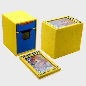 May include: A yellow plastic card storage box with a blue drawer and a matching yellow card display frame. The box and frame feature engraved line patterns and the text WANTED DEAD OR ALIVE. A card depicting an anime character is visible inside.