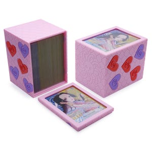 May include: A light pink plastic deck box with a textured scale pattern, featuring red and purple heart-shaped accents. The box includes a removable lid with a clear window displaying a trading card of a character labeled Hancock.