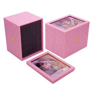 May include: A pink plastic card storage box with a floral pattern and a yellow center, featuring a clear window on the lid displaying a Nico Robin trading card. The set includes an open box filled with dark cards and a matching lid with a card.