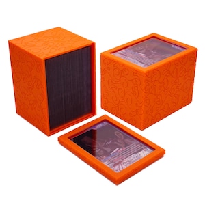May include: An orange plastic deck box for trading cards featuring a textured flame pattern on the exterior. The set includes one open box showing a stack of dark cards, one closed box with a clear top window, and one matching orange frame.