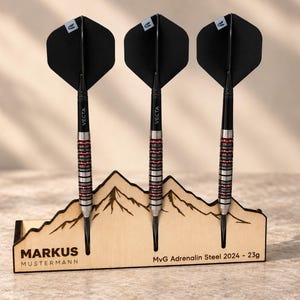 Op de afbeelding: Drie MvG Adrenalin Steel 2024 23g darts met zwarte flights en zilveren en rode barrels, gepresenteerd in een lichte houten standaard in de vorm van een bergketen. De standaard bevat de tekst MARKUS MUSTERMANN en MvG Adrenalin Steel 2024 - 23g.