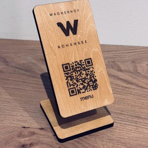 Könnte beinhalten: Ein rechteckiger Aufsteller aus hellem Holz mit einer vertikalen Anzeigetafel, die den schwarzen Schriftzug WAGNERHOF, einen großen Buchstaben W, den Text ACHENSEE, einen schwarzen QR-Code und das Wort menu auf der Holzoberfläche zeigt.