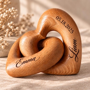 May include: Two interlocking wooden hearts with a smooth, light brown wood grain finish. The surface is engraved with the name Emma on the lower heart, and the name Liam and the date 05.18.2025 on the upper heart in a dark, elegant script font.