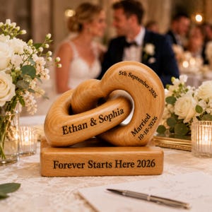 May include: A light-coloured wooden sculpture featuring two interlocking heart-shaped links resting on a rectangular base. The wood is engraved with the text Ethan & Sophia, 5th September 2026, Married 10-18-2026, and Forever Starts Here 2026.