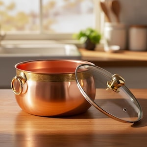 May include: A round copper pot with a polished finish, featuring a gold-toned band with a Greek key pattern around the rim. It has two gold-toned ring handles on the sides and comes with a clear glass lid topped with a gold-toned knob handle.
