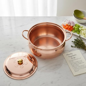 May include: A polished copper pot with two side handles and a matching lid featuring a decorative embossed pattern and a gold-toned knob. Beside the pot lies a recipe card titled Hearty Vegetable Soup, alongside chopped vegetables and herbs.